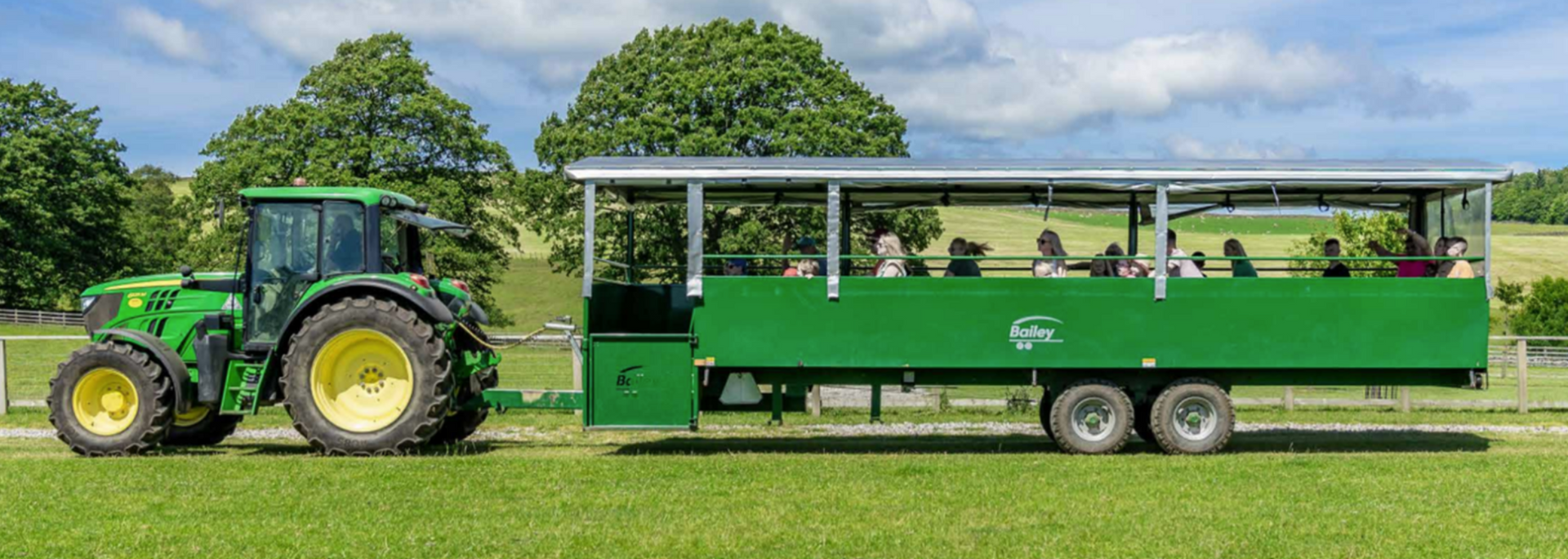 A green tractor pulling a green open-air trailer with people in it, driving on grass, with trees in the background.