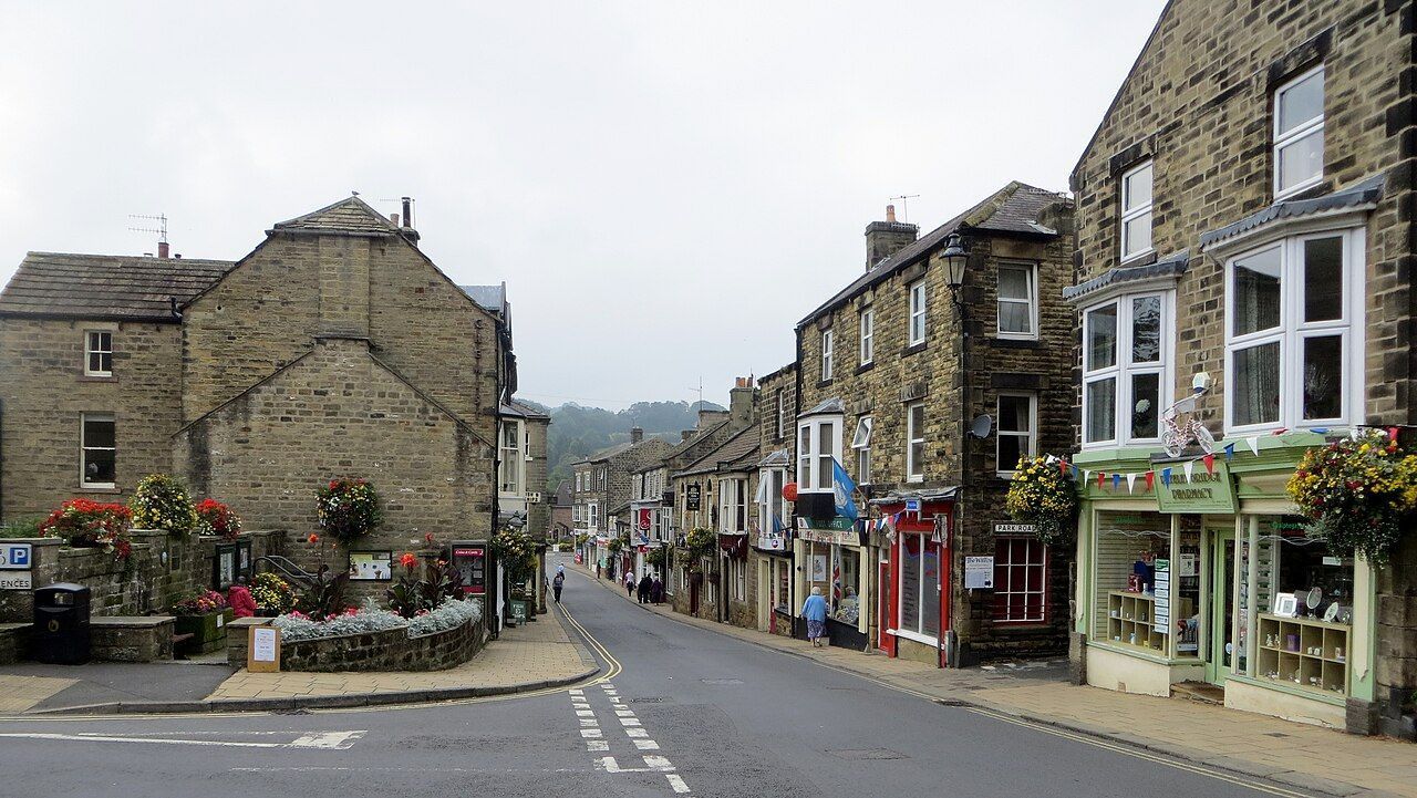 Street scene with stone buildings, shops, and a road curving downhill. Cloudy sky.