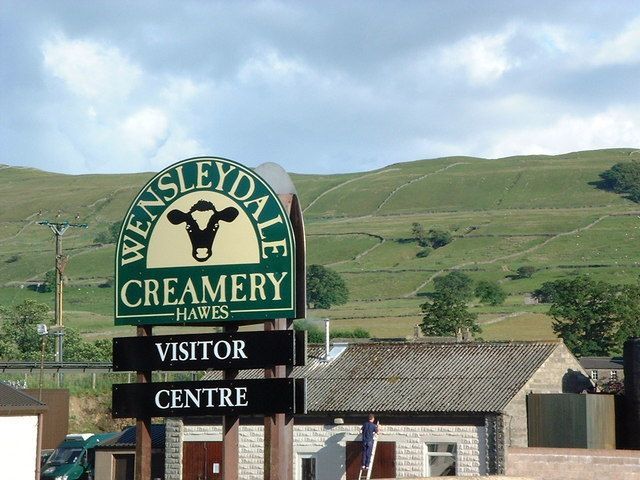 Sign for Wensleydale Creamery, Hawes, with a green background and a cow image, beside a stone building and hills.