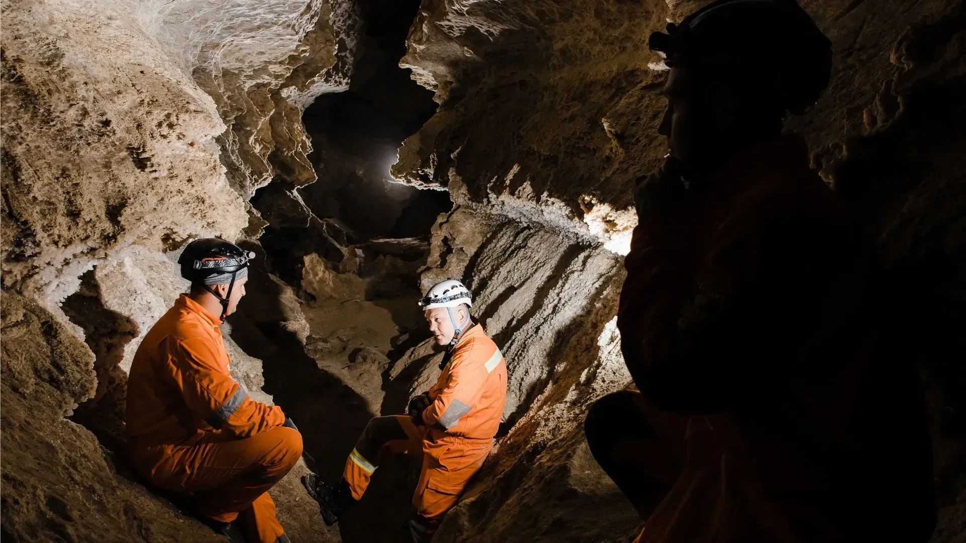 Three people in orange suits and helmets explore a dark cave, illuminated by headlamps.