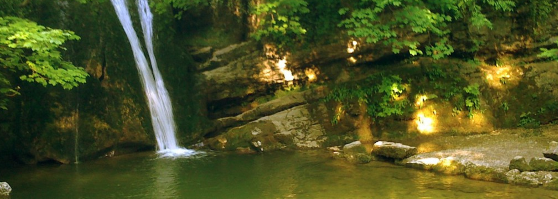 Waterfall cascading into a calm green pool, surrounded by lush green foliage and sunlit rocks.