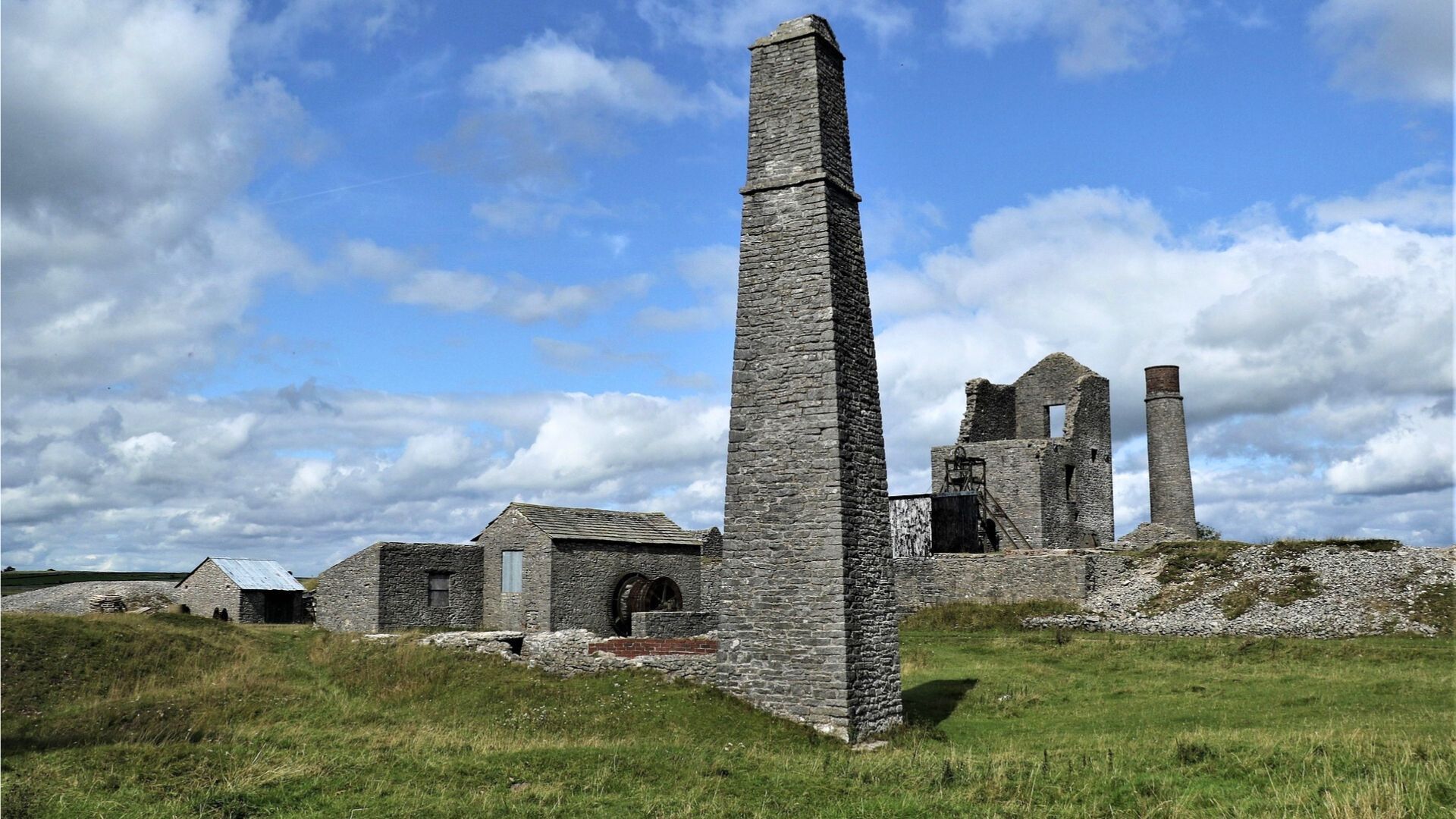 Ruined stone buildings with a tall, narrow chimney against a blue sky with clouds.