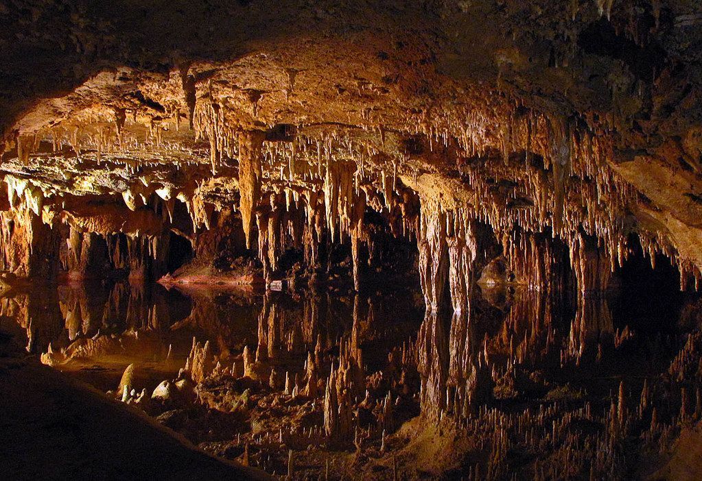 Cave interior with stalactites and reflections in water, illuminated by warm light.