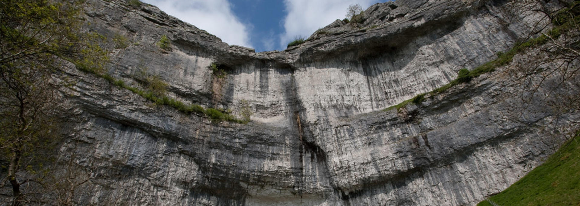 A dramatic layered rock formation, with green patches, viewed from below under a blue and cloudy sky.