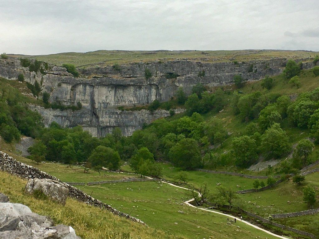 Limestone gorge with lush green trees and grassy valley, overcast sky.