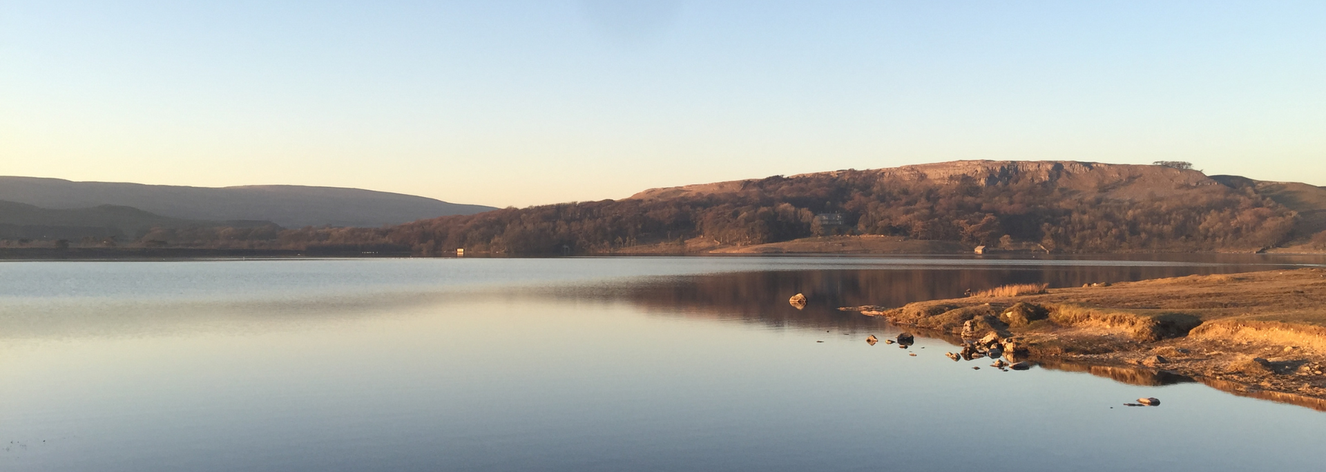 Calm lake reflects a forested hillside under a clear blue sky.