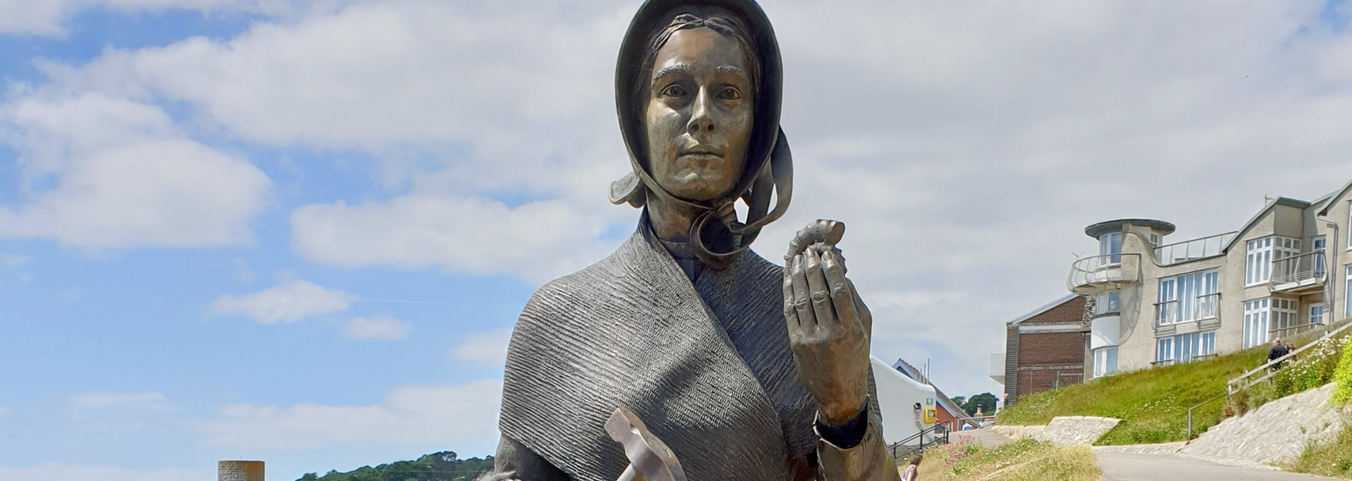 Statue of a woman holding something, set by the water with buildings and a cloudy sky.