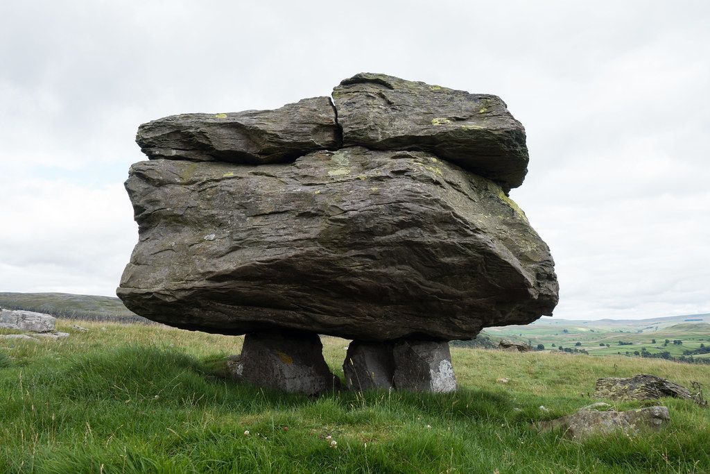 Large, weathered rock formation balanced on smaller stone supports in a grassy field, against a cloudy sky.