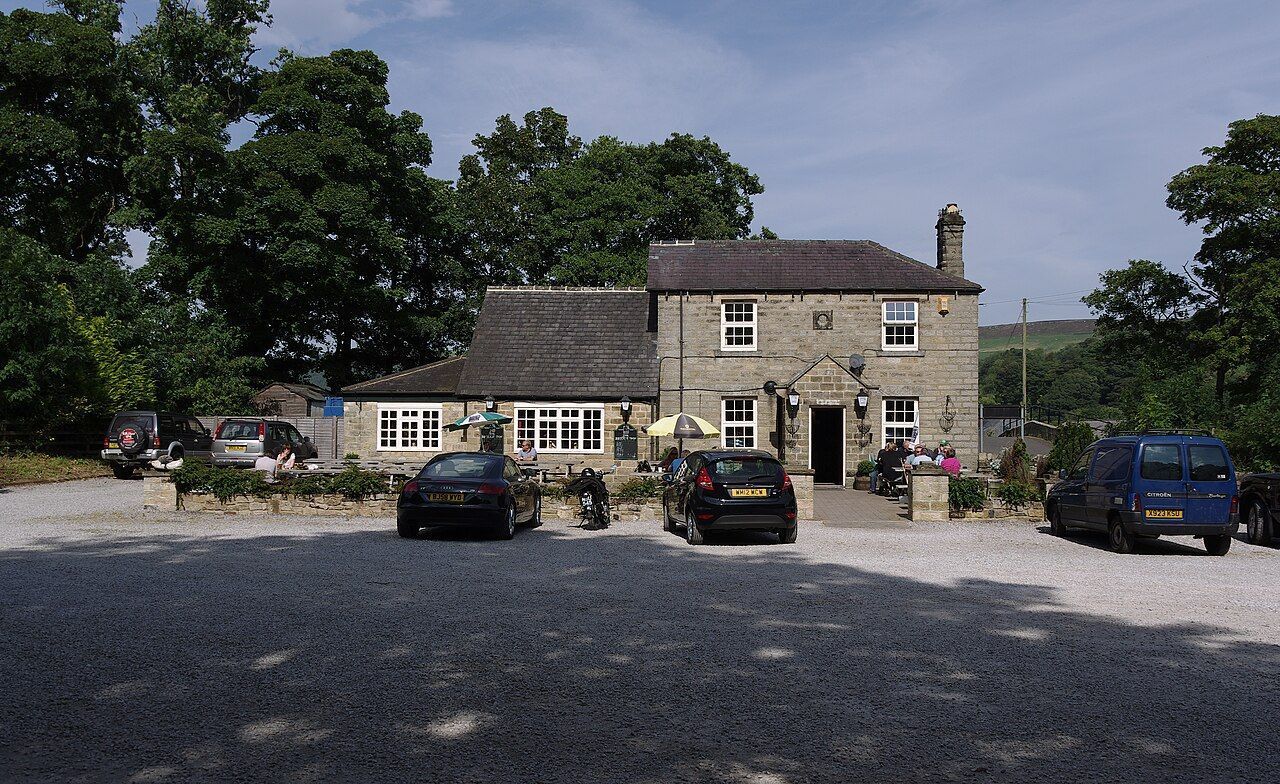 Stone pub with gravel parking area; cars parked in front. Trees in background.