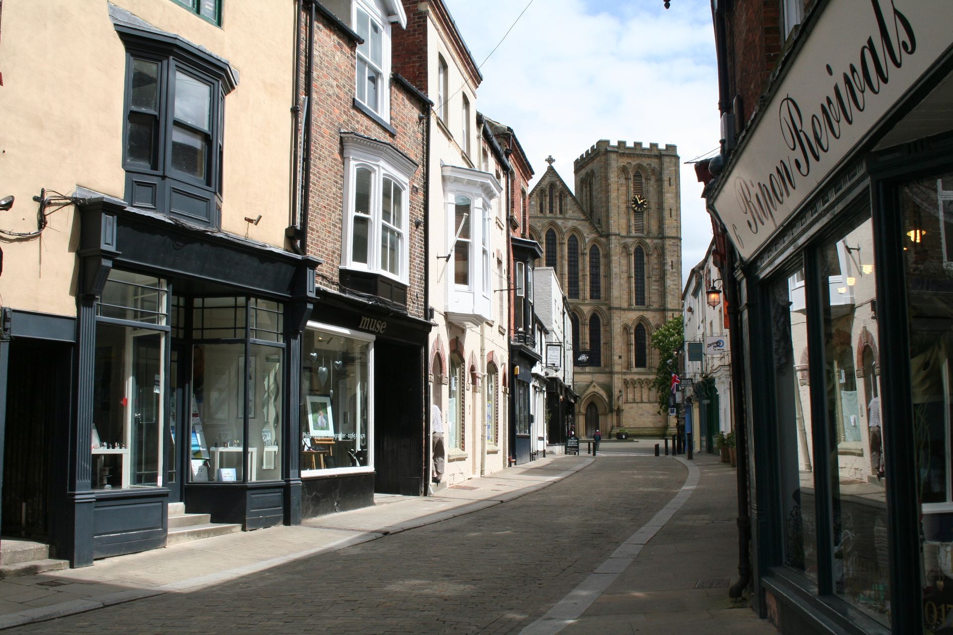 Street lined with shops, leading to a tall church tower. Beige and black shopfronts. Sunny day.