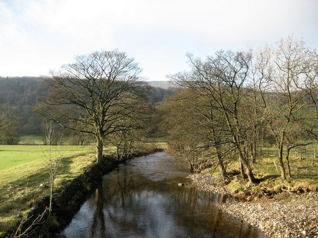A river flowing between grassy banks, lined with bare trees, under a cloudy sky.