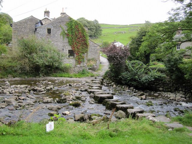 Stone house beside a shallow river with stepping stones. Lush greenery and cloudy sky.