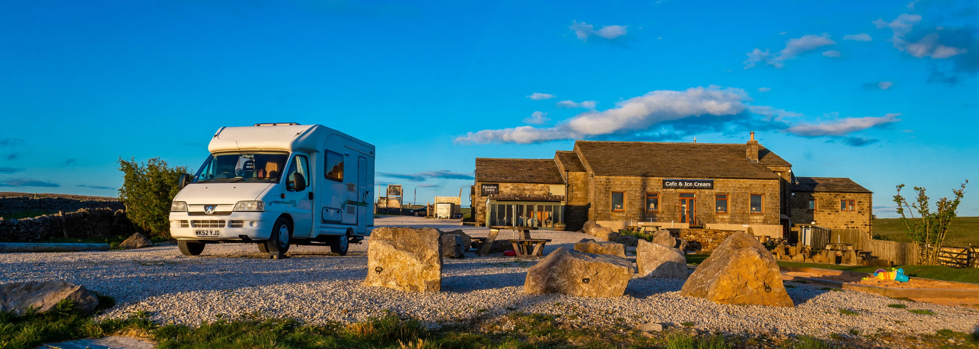 A white RV on a gravel road, approaching a stone building under a bright blue sky.