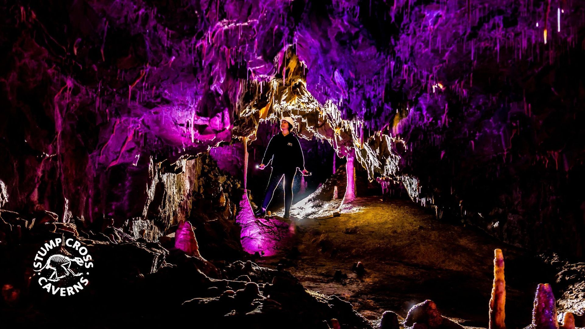 Person in a cave lit with purple and yellow lights, surrounded by stalactites and stalagmites.