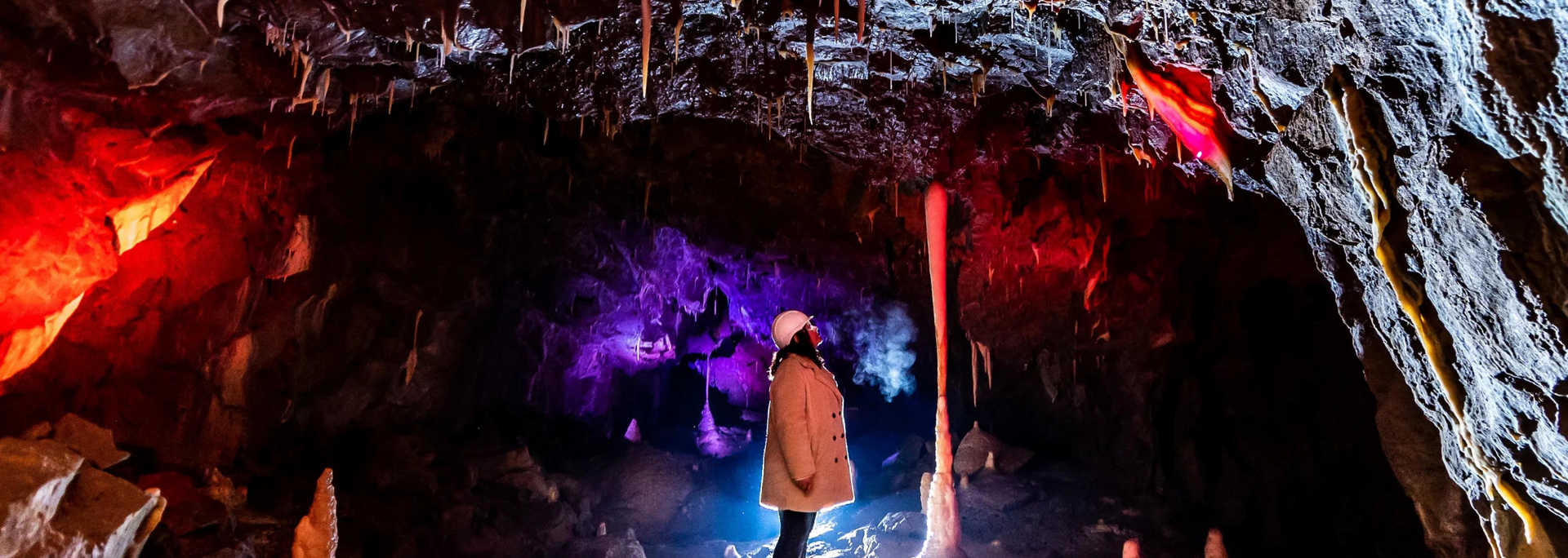 Person in a cave lit with colored lights. Stalactites hang from the cave ceiling.