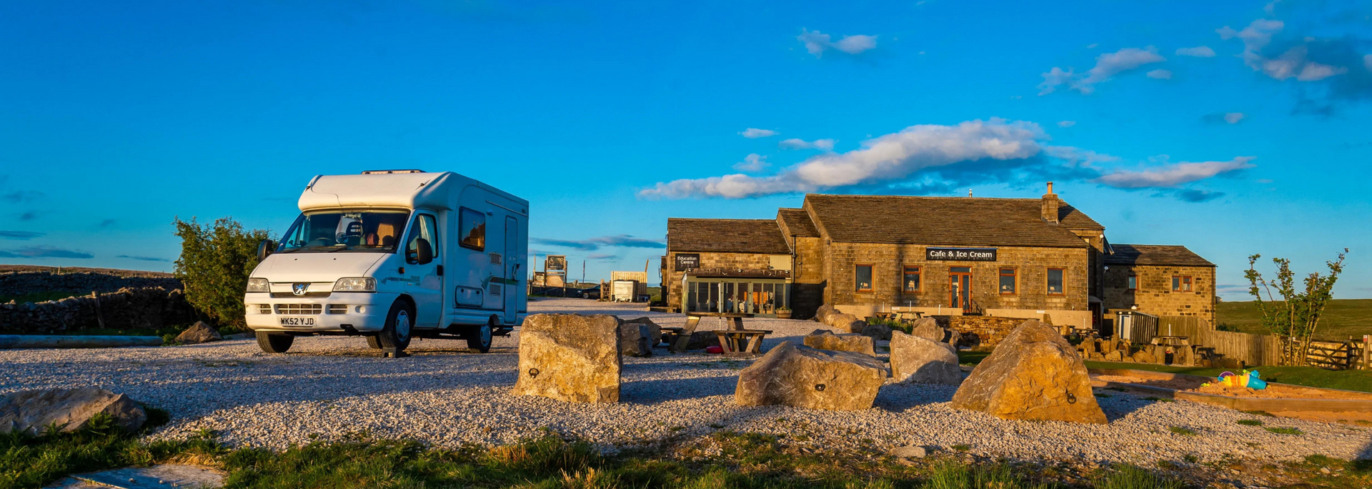 White campervan on a gravel road, passing a stone building on a sunny day with blue sky.