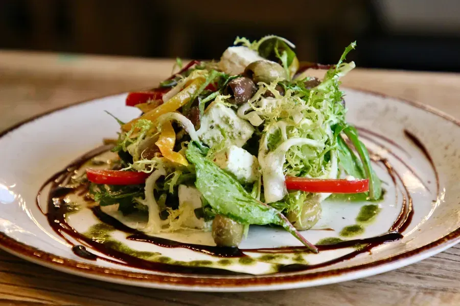 Salad on a plate, featuring greens, olives, peppers, and cheese with drizzle.