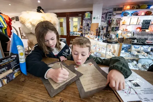 Two children write on aged paper at a shop counter, focused. The boy has a surprised expression.
