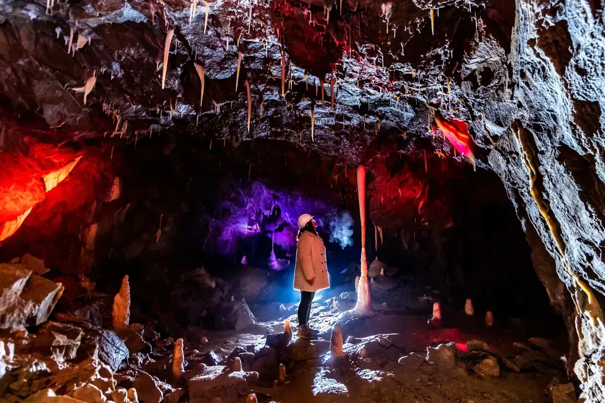 Person standing in a cave with colored lights illuminating stalactites and stalagmites.