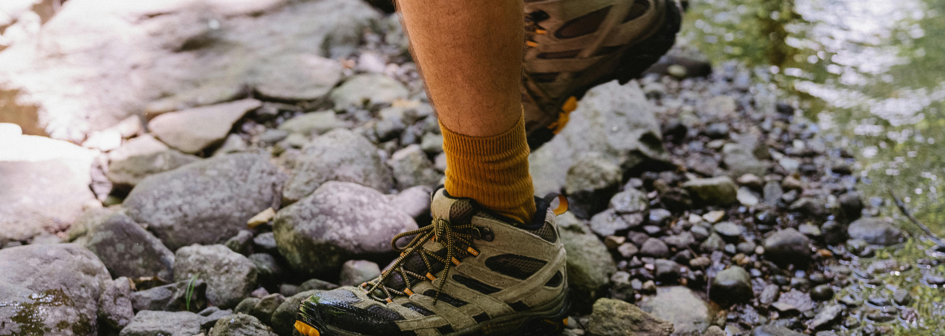 A person's leg and hiking boot stepping on a rocky path with a water stream to the right.