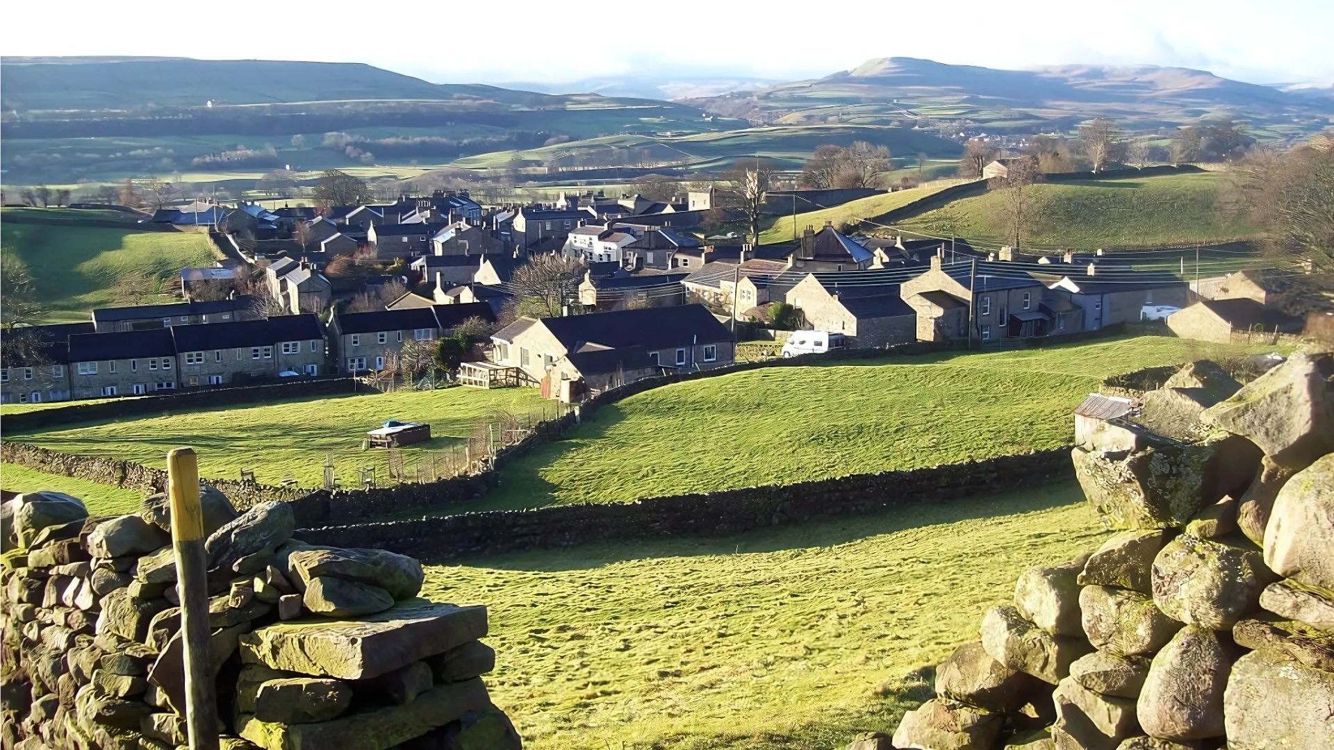 View of a village nestled in a valley, green fields, stone walls in foreground, hills in the background.