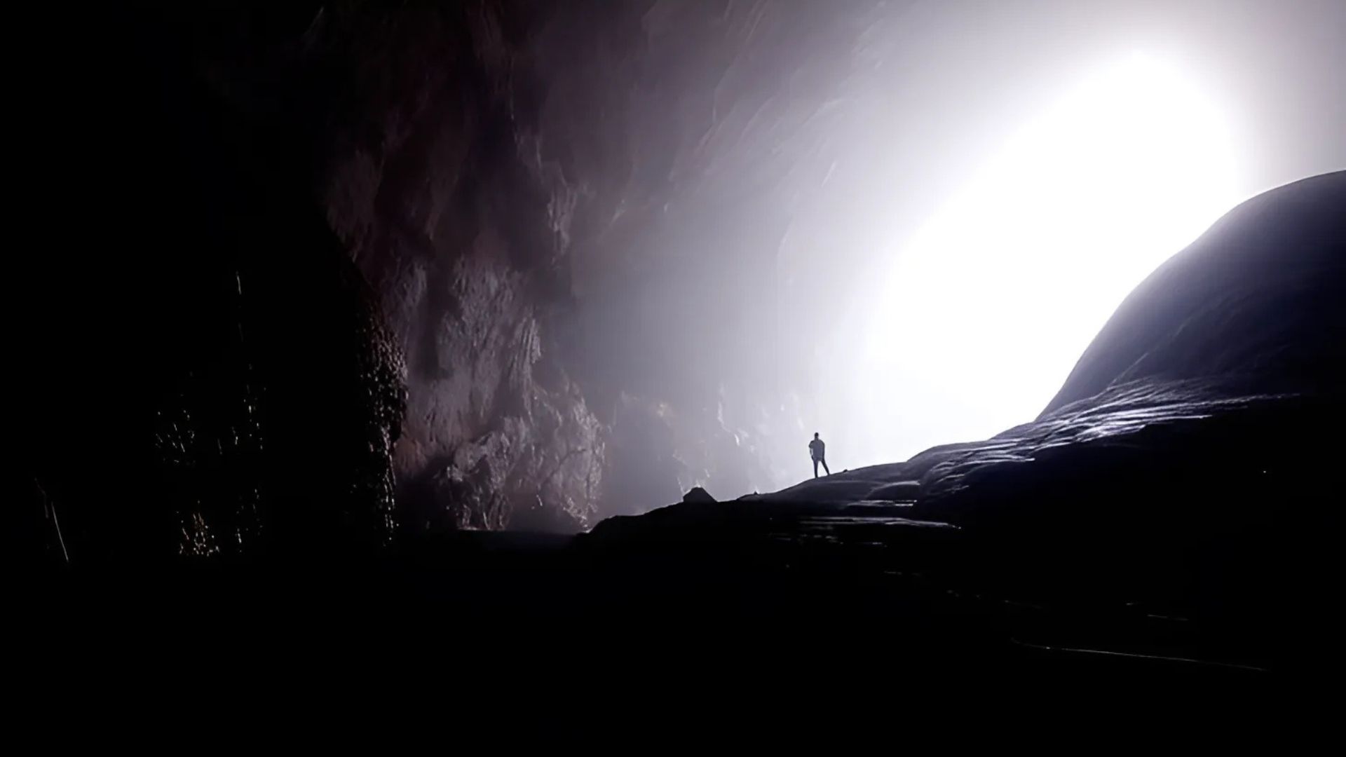 Silhouette of a person standing at a cave entrance, looking out towards a bright light.
