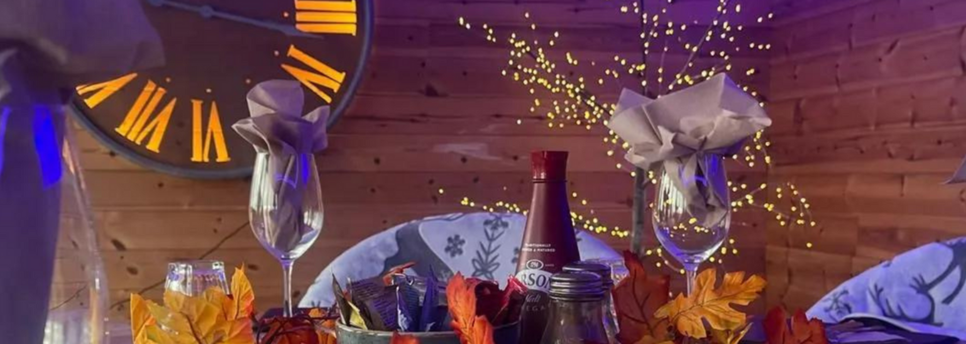 Table setting with champagne flutes, a bottle of wine, and fall foliage decorations, in front of a rustic clock.