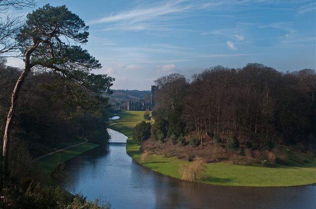 A wide river winds through a green meadow surrounded by trees, with a castle in the distance under a blue sky.