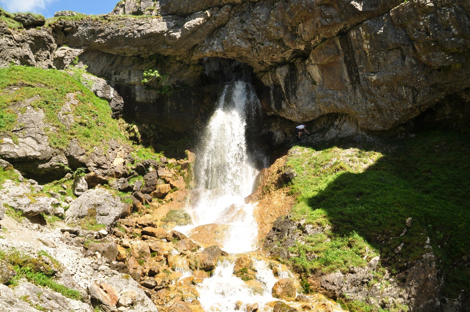 Waterfall cascading from a rocky overhang, surrounded by green grass and rocks.