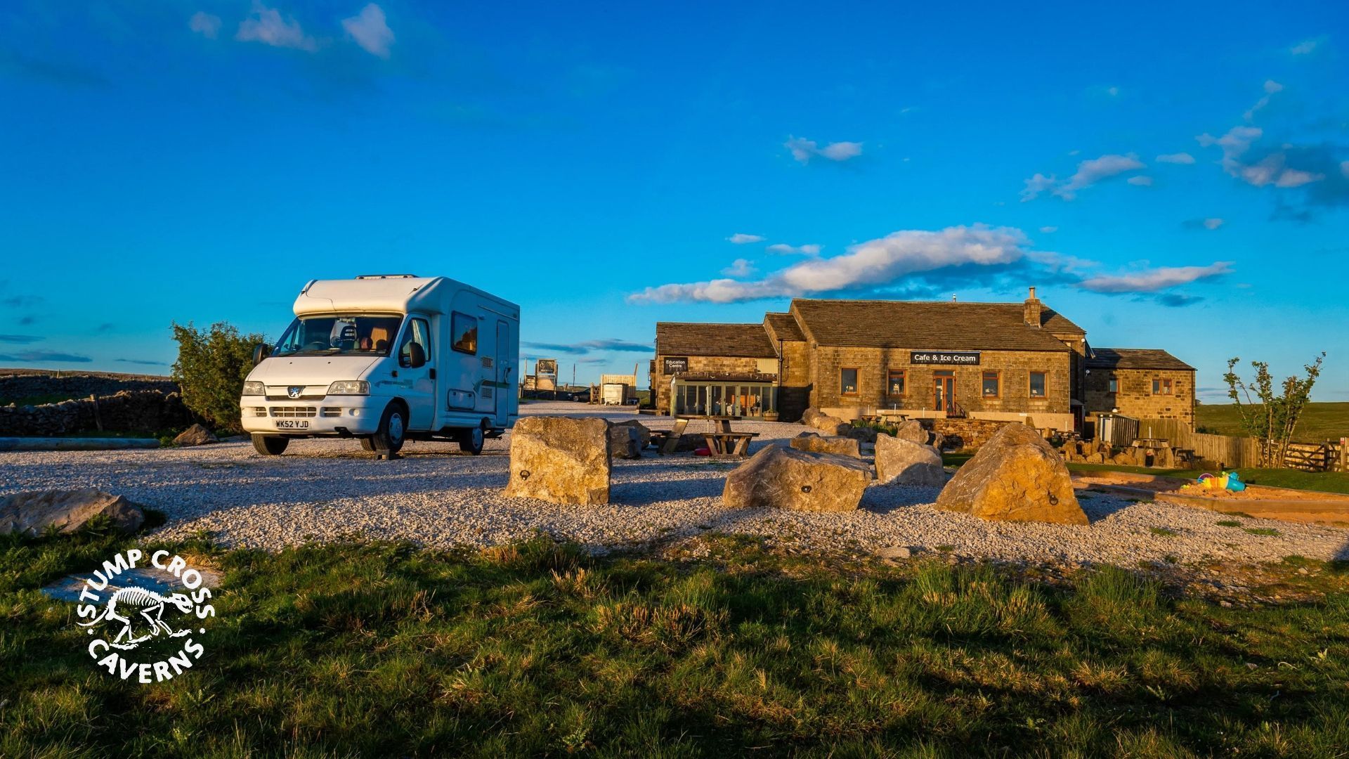 Motorhome parked on a gravel drive near a stone building under a blue sky, grass in foreground.