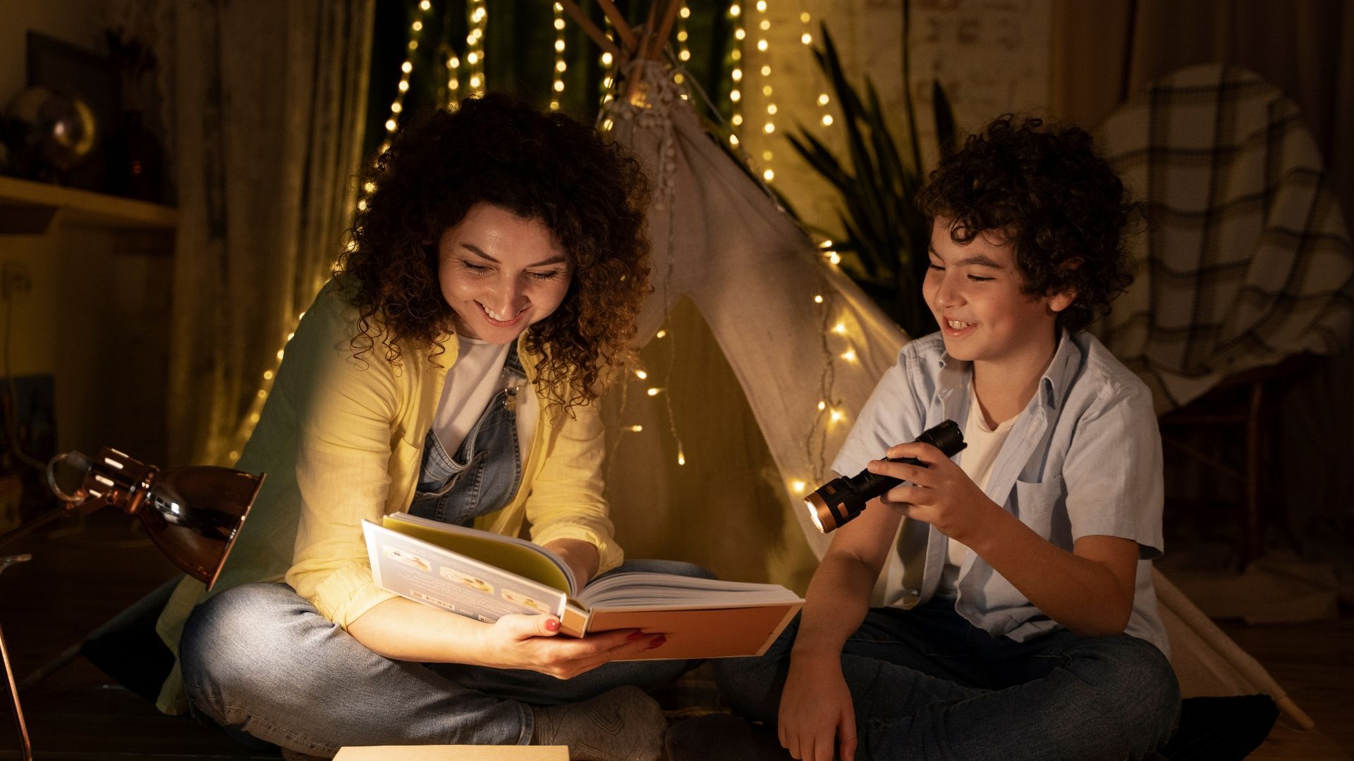 Woman and child reading a book inside a tent lit by fairy lights. Child holds a flashlight.
