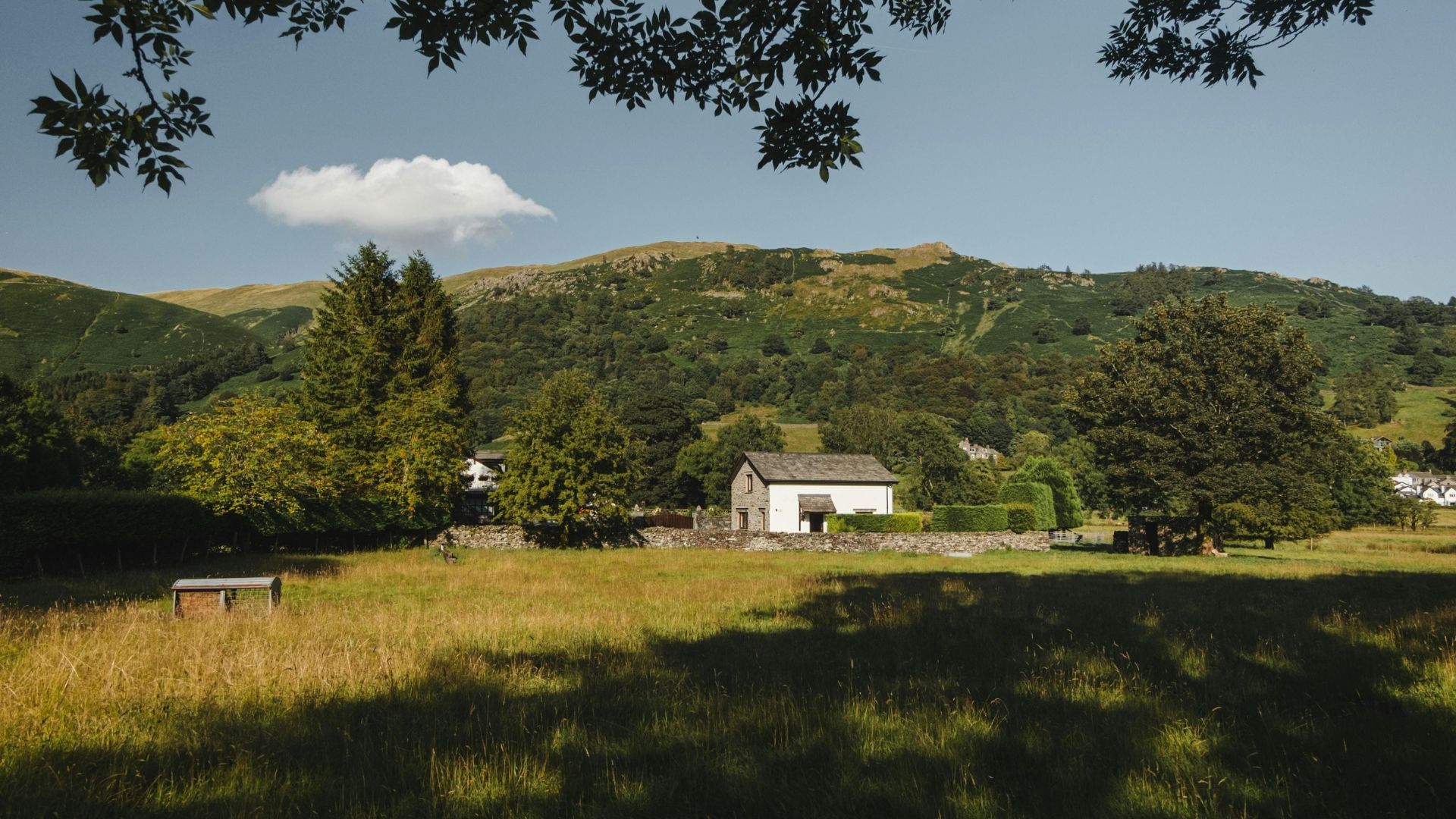 White building nestled in a valley with rolling green hills and a blue sky; sunlight through trees in the foreground.