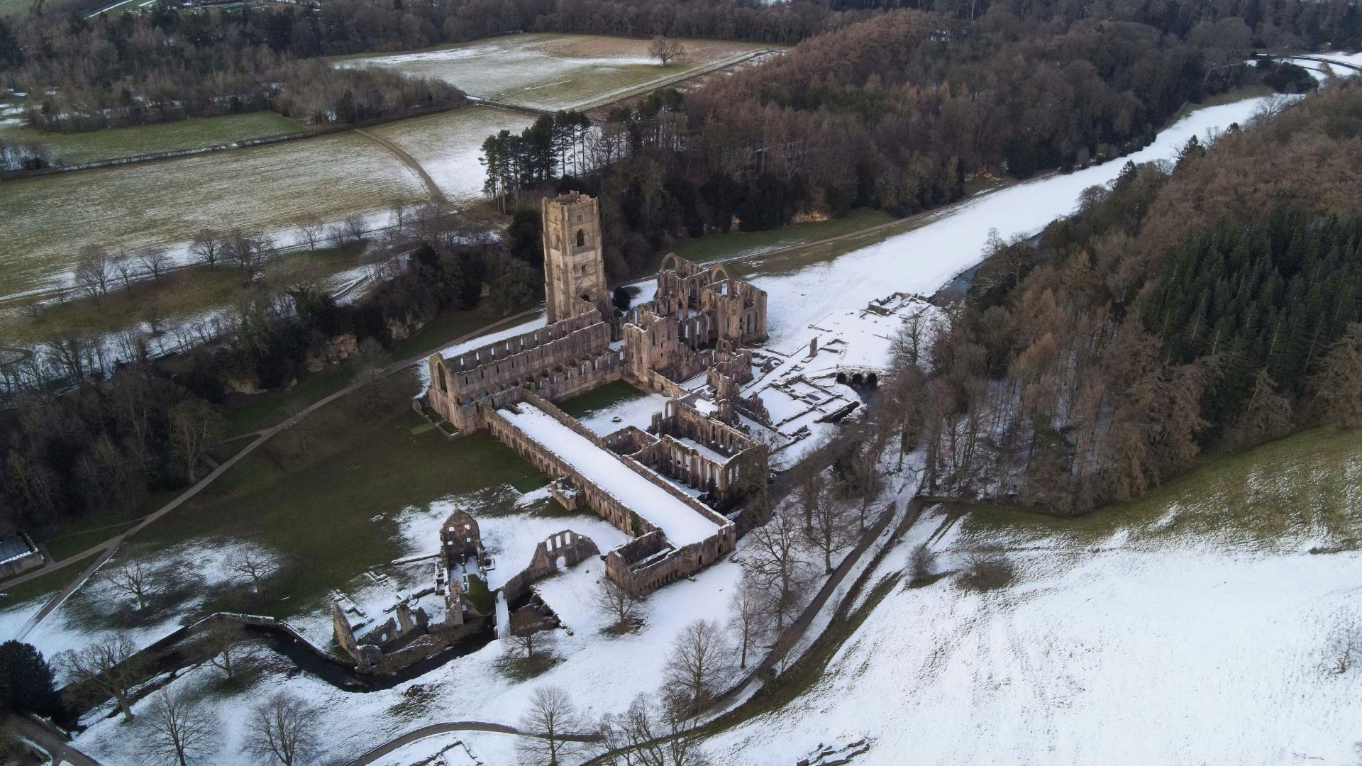 Aerial view of Fountains Abbey ruins in winter, covered in snow, near a river and forest.