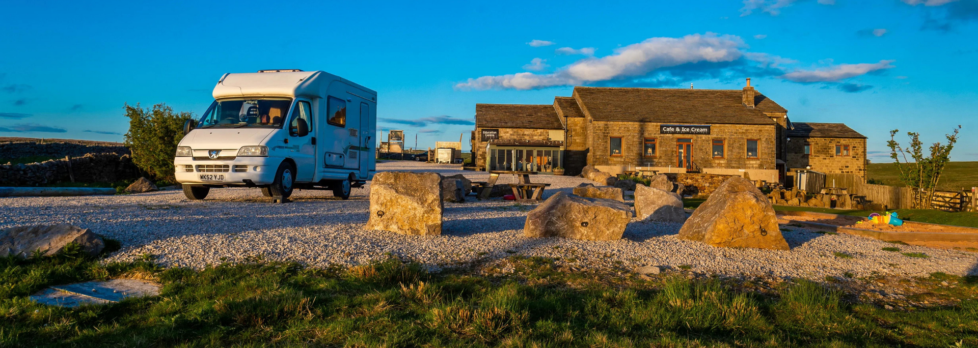 White RV on a gravel road, approaching a stone building under a blue sky.