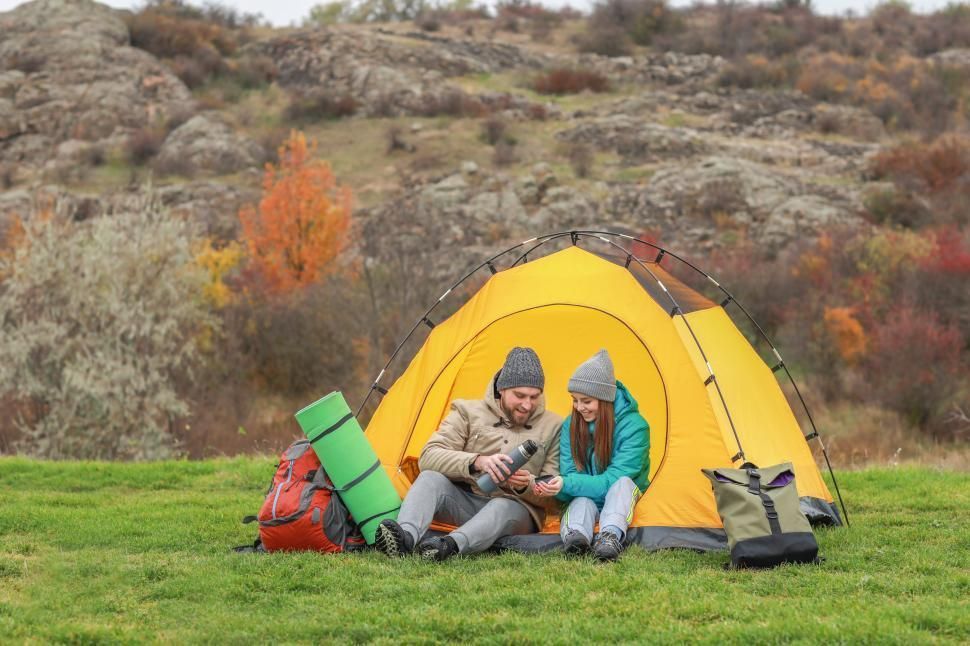 Couple in hats, sits outside a yellow tent on a grassy hill; they hold a thermos, with fall foliage in background.