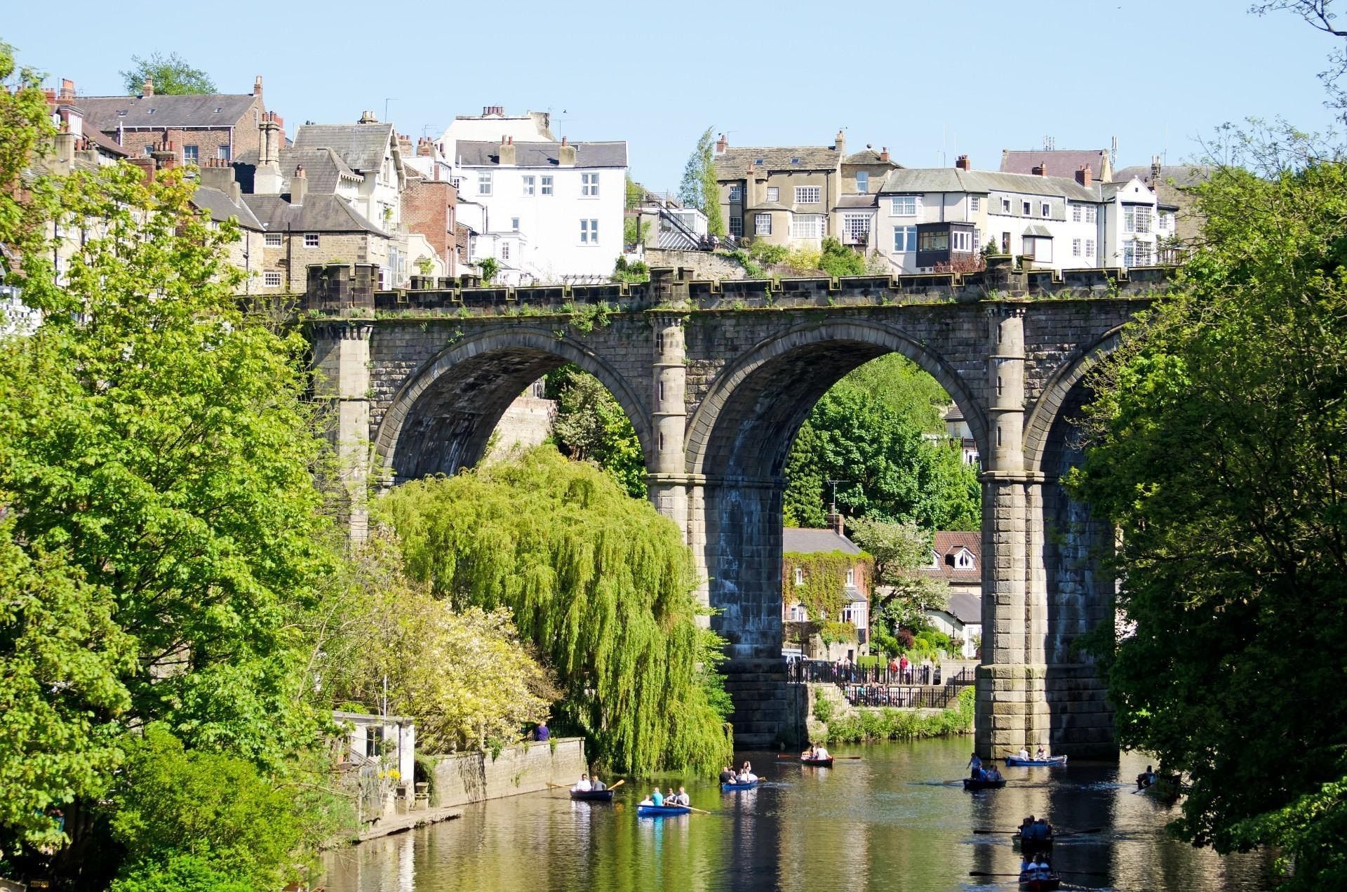 Stone viaduct bridge over a river with several boats. Buildings on a hill in the background, surrounded by trees.