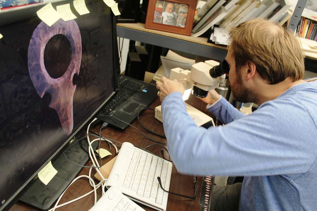 Man looking into a microscope, examining a graphic on a computer screen. Workspace with monitor, keyboard.
