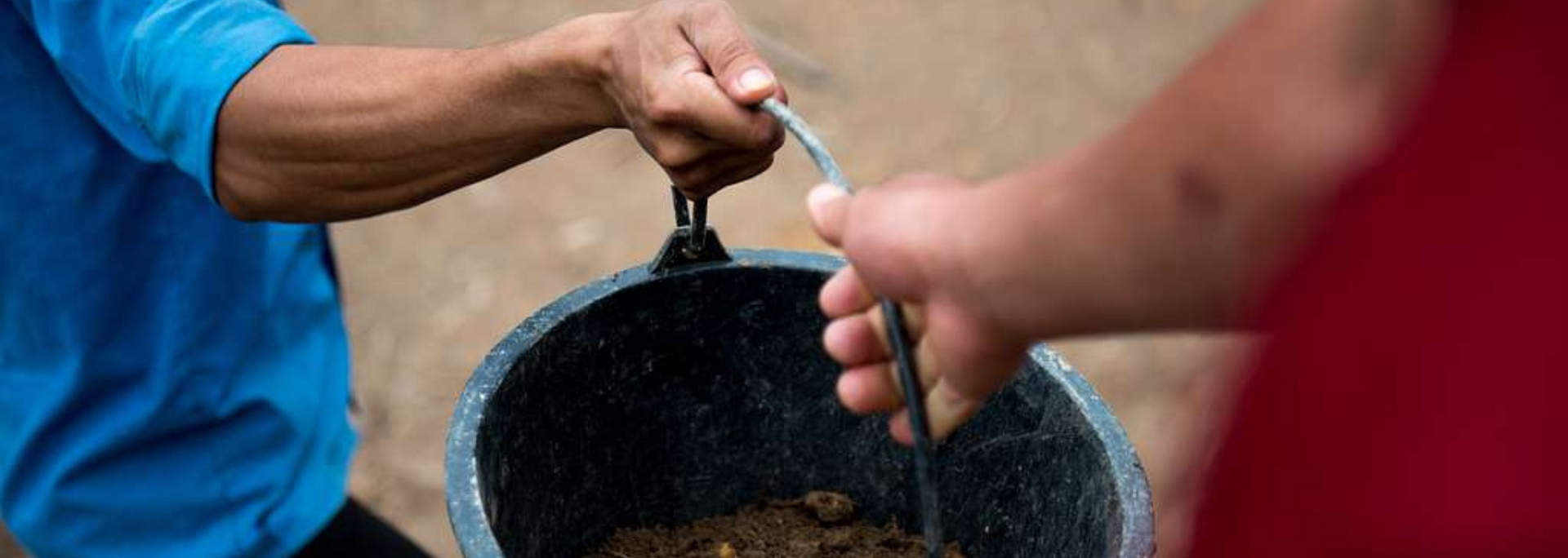 Two people holding a bucket of brown contents, one in blue, the other in red.