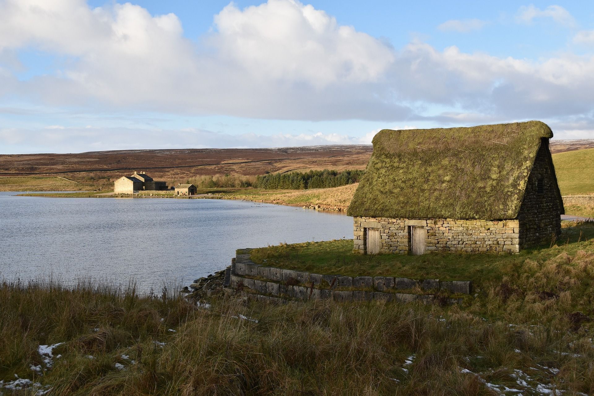 Stone hut with thatched roof beside a lake, rolling hills in the background under a cloudy sky.