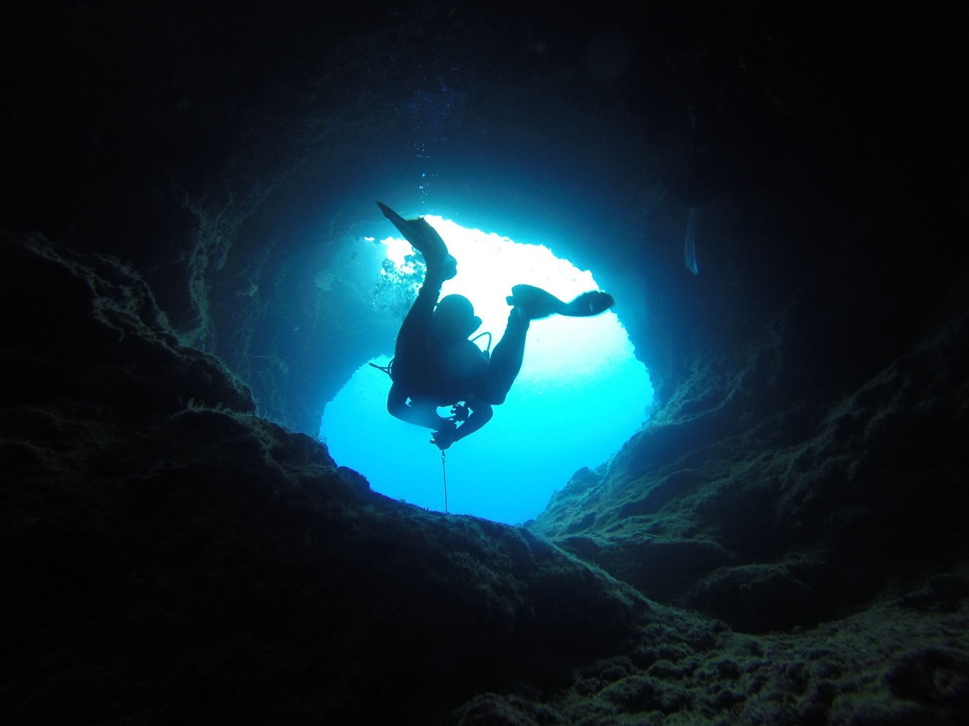 Diver in silhouette exits an underwater cave, illuminated by bright blue sunlight.