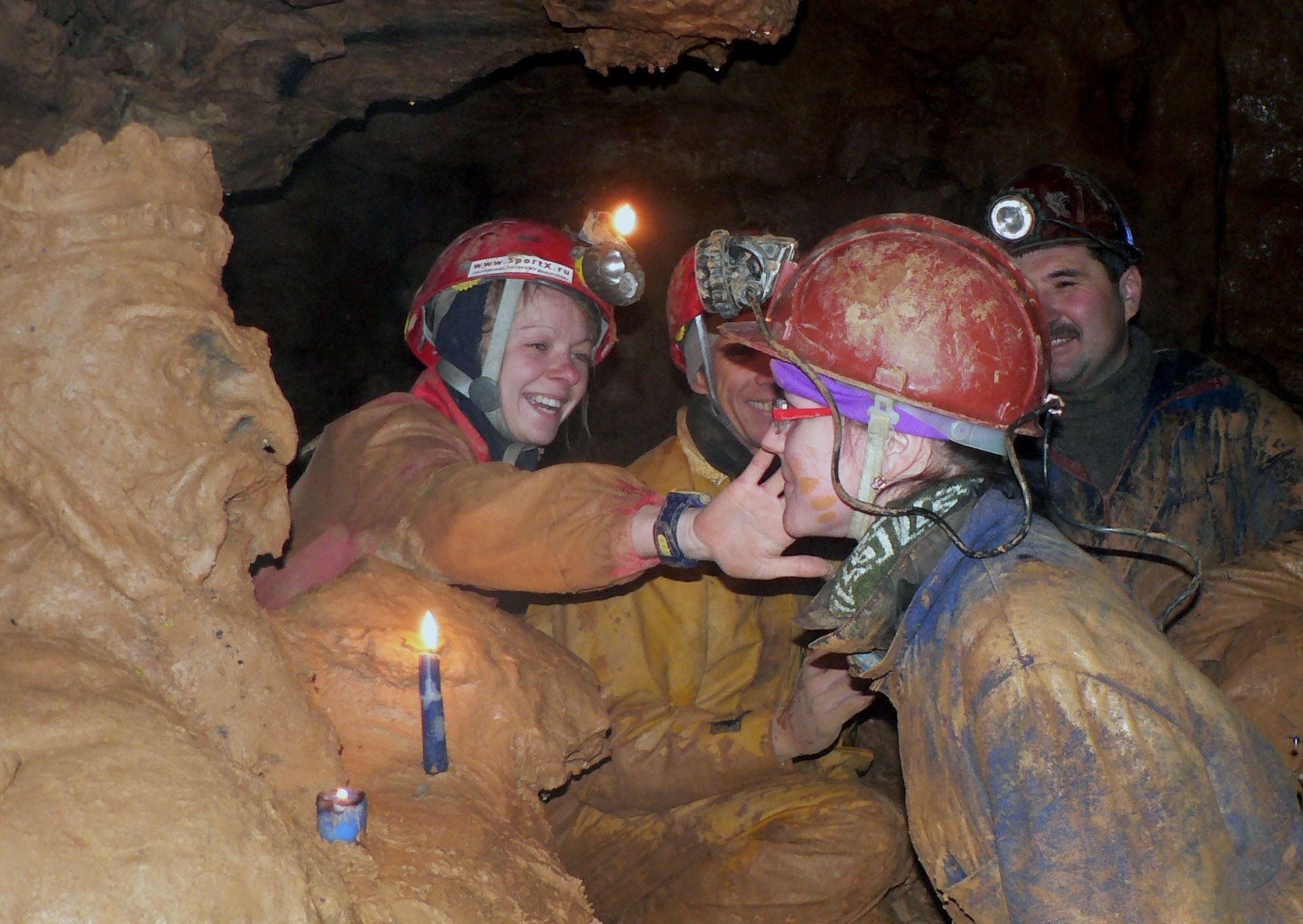 People in cave with headlamps and helmets, covered in mud. One person smiles, touching another's nose. Candles illuminate the scene.