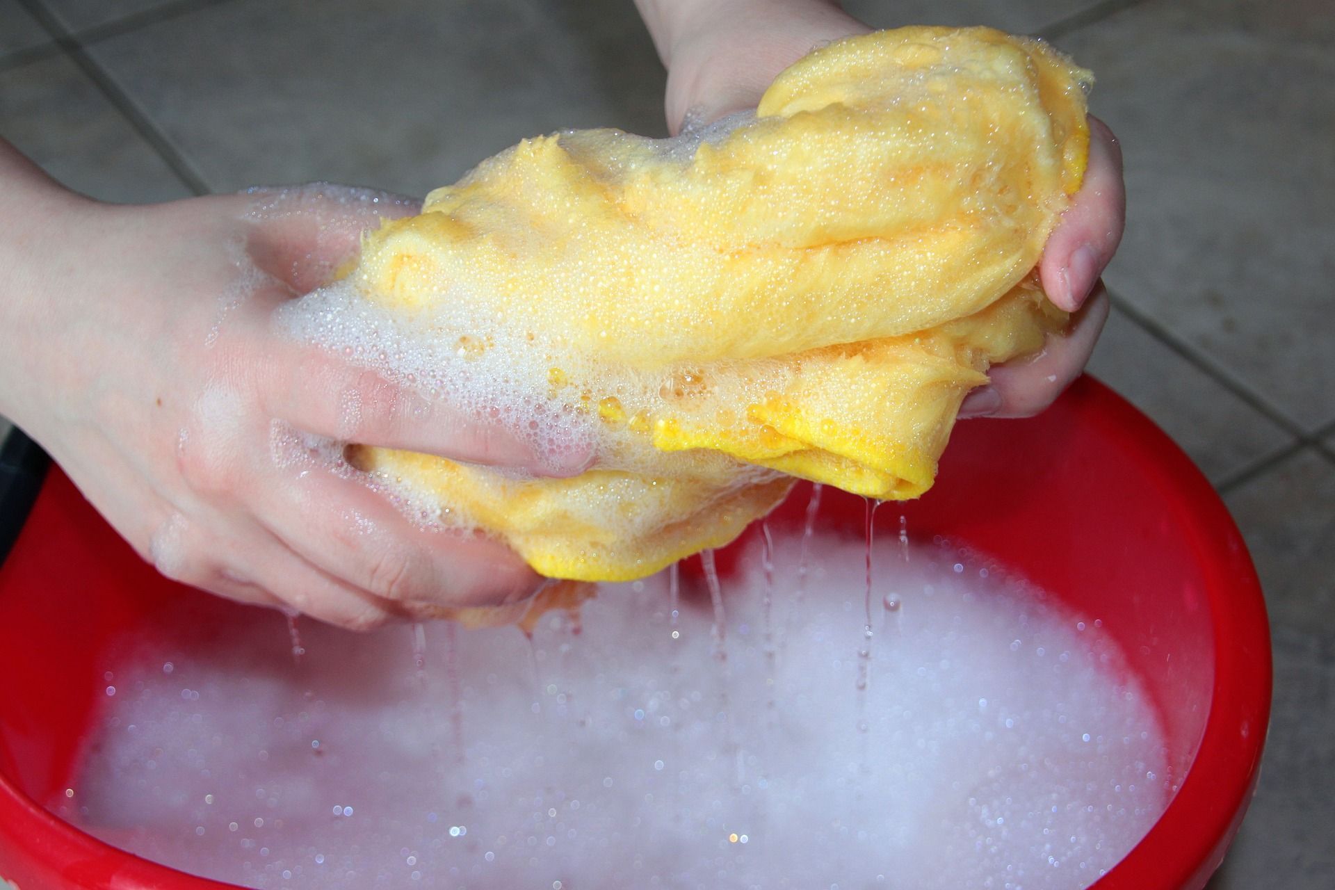 Hands squeezing a yellow sponge in soapy water inside a red bucket.