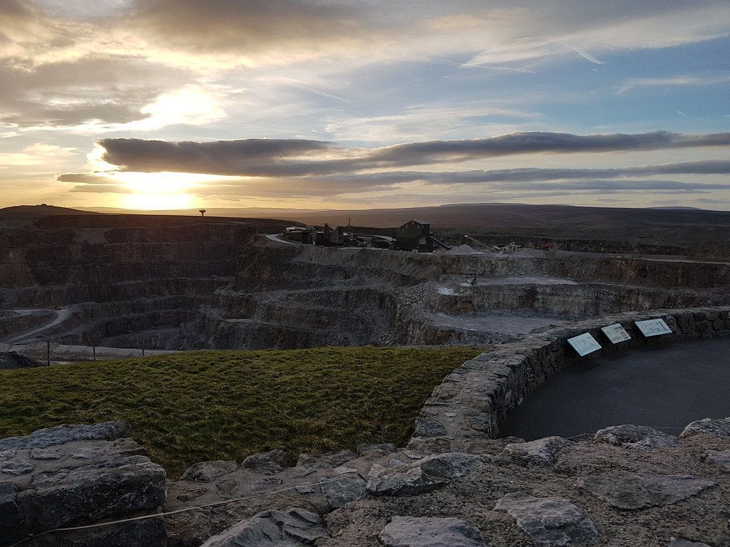 Sunset view over a quarry, with a stone wall in the foreground. Sky is cloudy, with the sun low on the horizon.