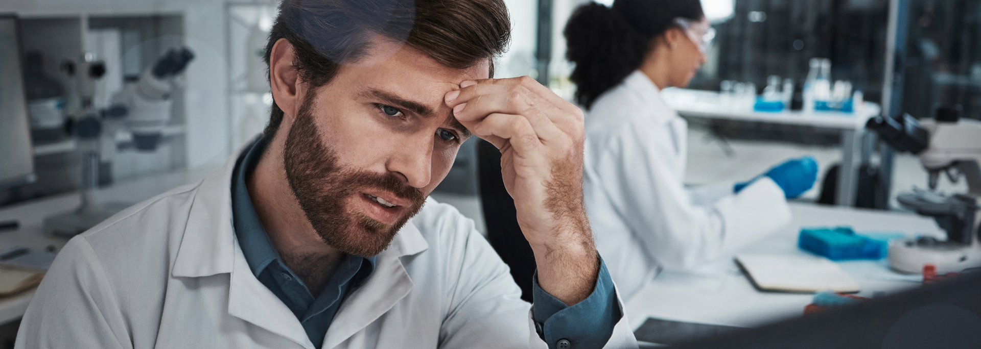 A scientist in a lab coat looks stressed, holding his forehead. Another scientist works nearby.