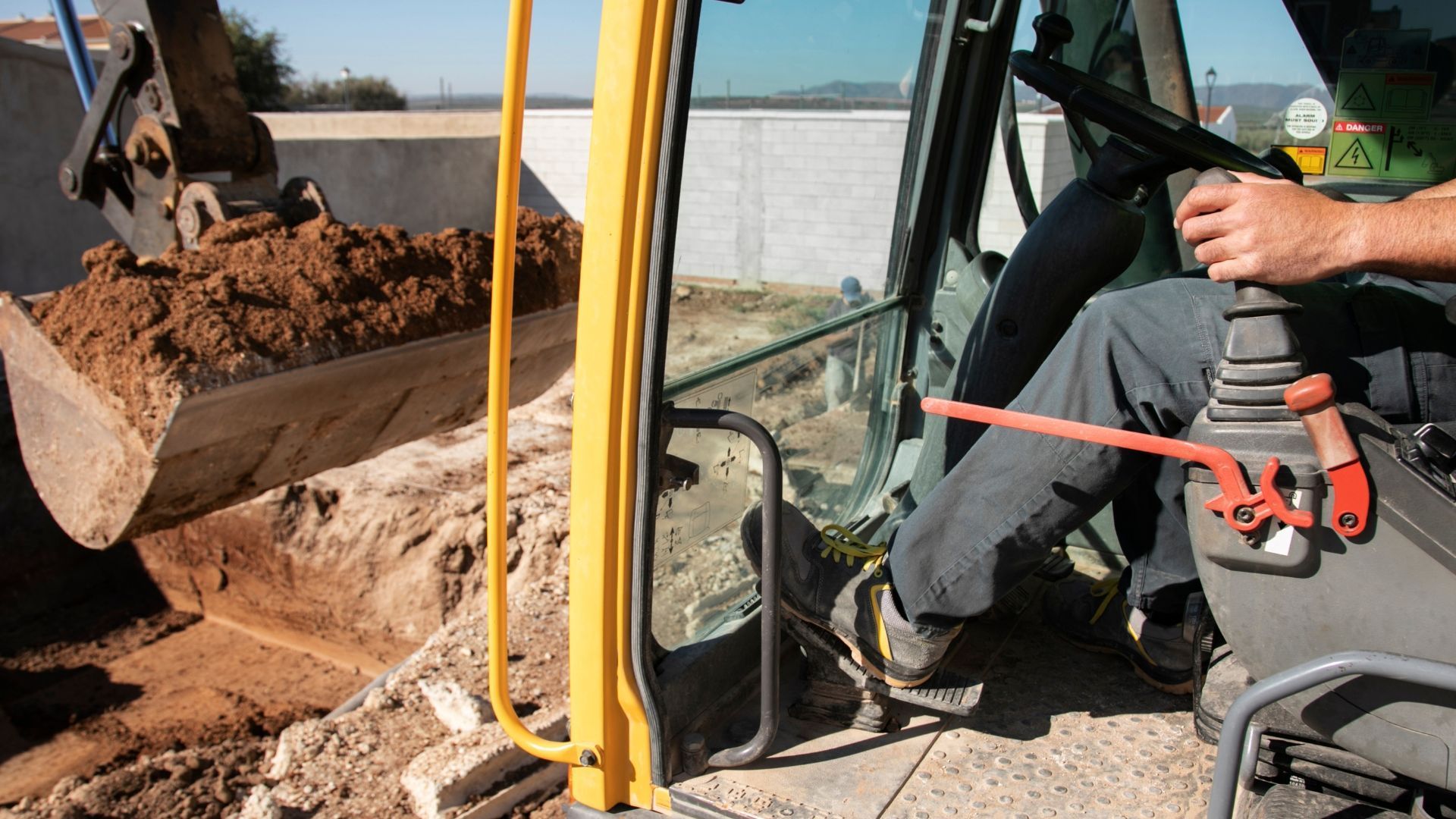 A person operating a yellow excavator, scooping dirt. The person's hand is on the controls.