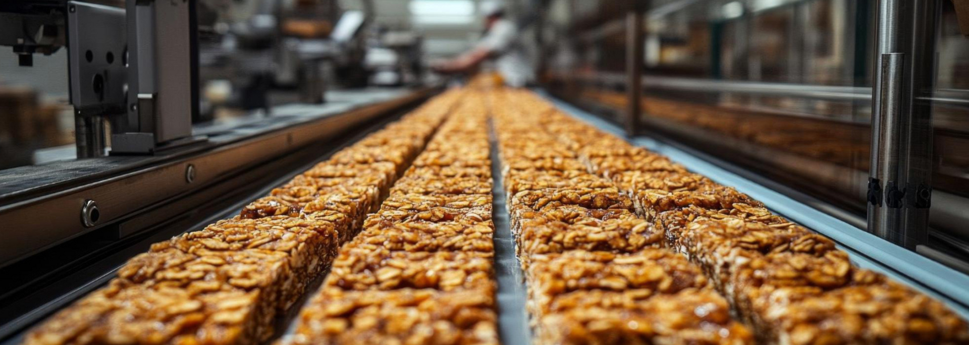 Granola bars on a factory conveyor belt, with a blurred person working in the background.