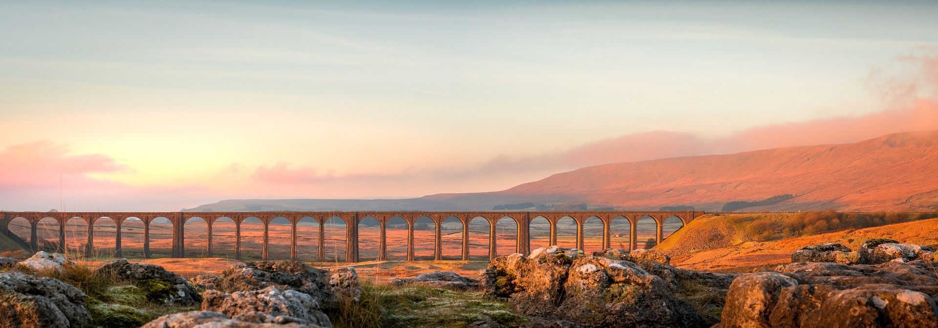 Long stone railway bridge against a colorful sunset, with rolling hills.