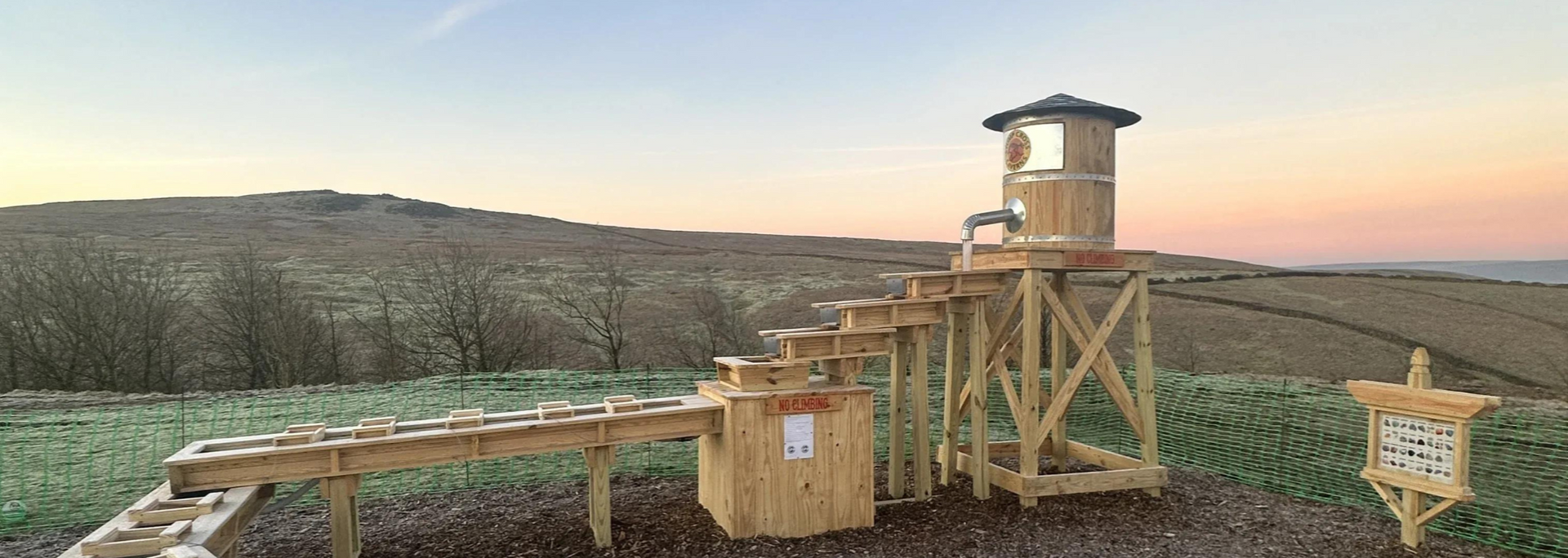Wooden structure resembling a water wheel or flume in a field, under a dusk sky.