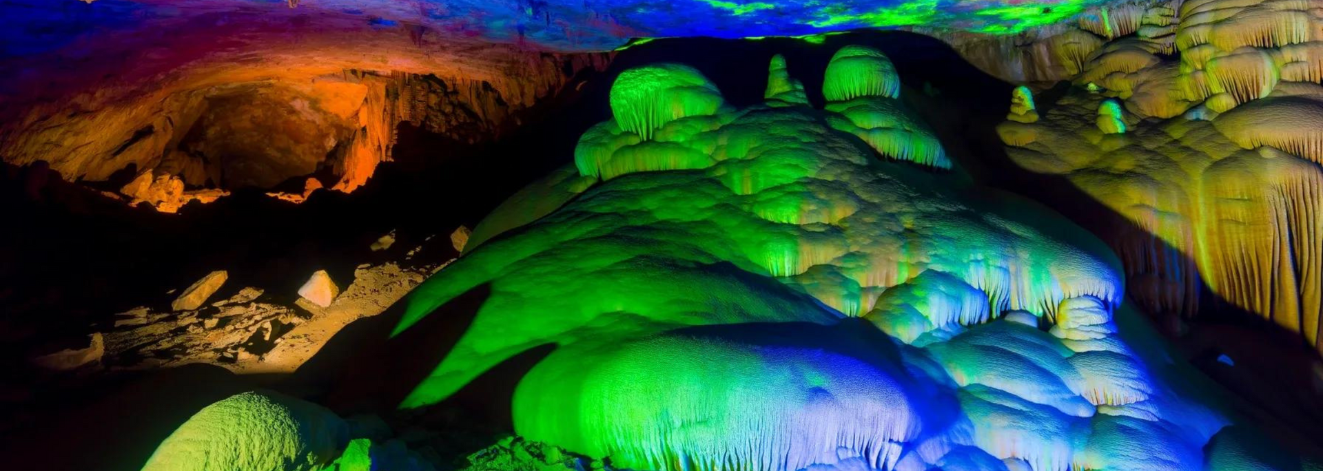 A cave interior illuminated with colorful lights. Green stalagmites and stalactites with orange and blue hues.