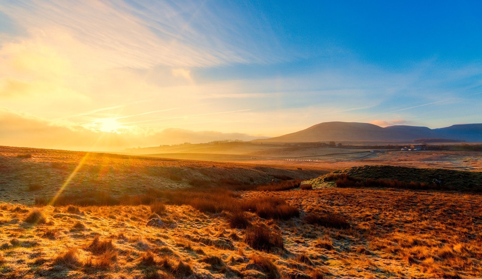 Sunrise over a dry, hilly landscape with mountains in the background, sky transitioning from orange to blue.